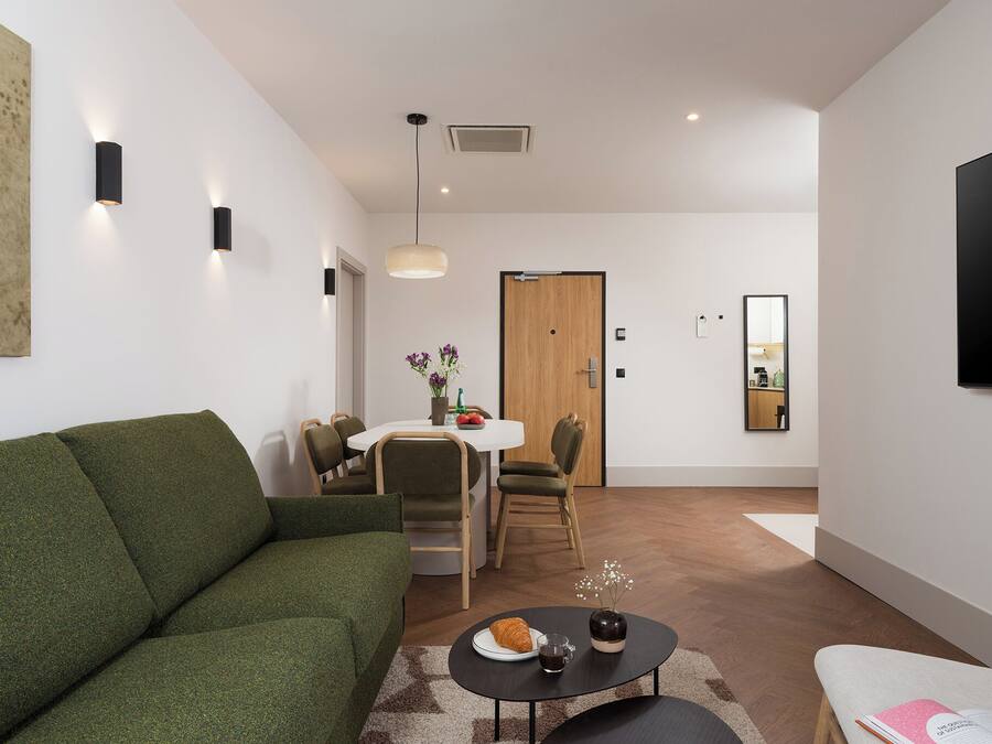 Una sala moderna con un sofá verde, un comedor con mesa y sillas blancas, una puerta de madera y un televisor montado.