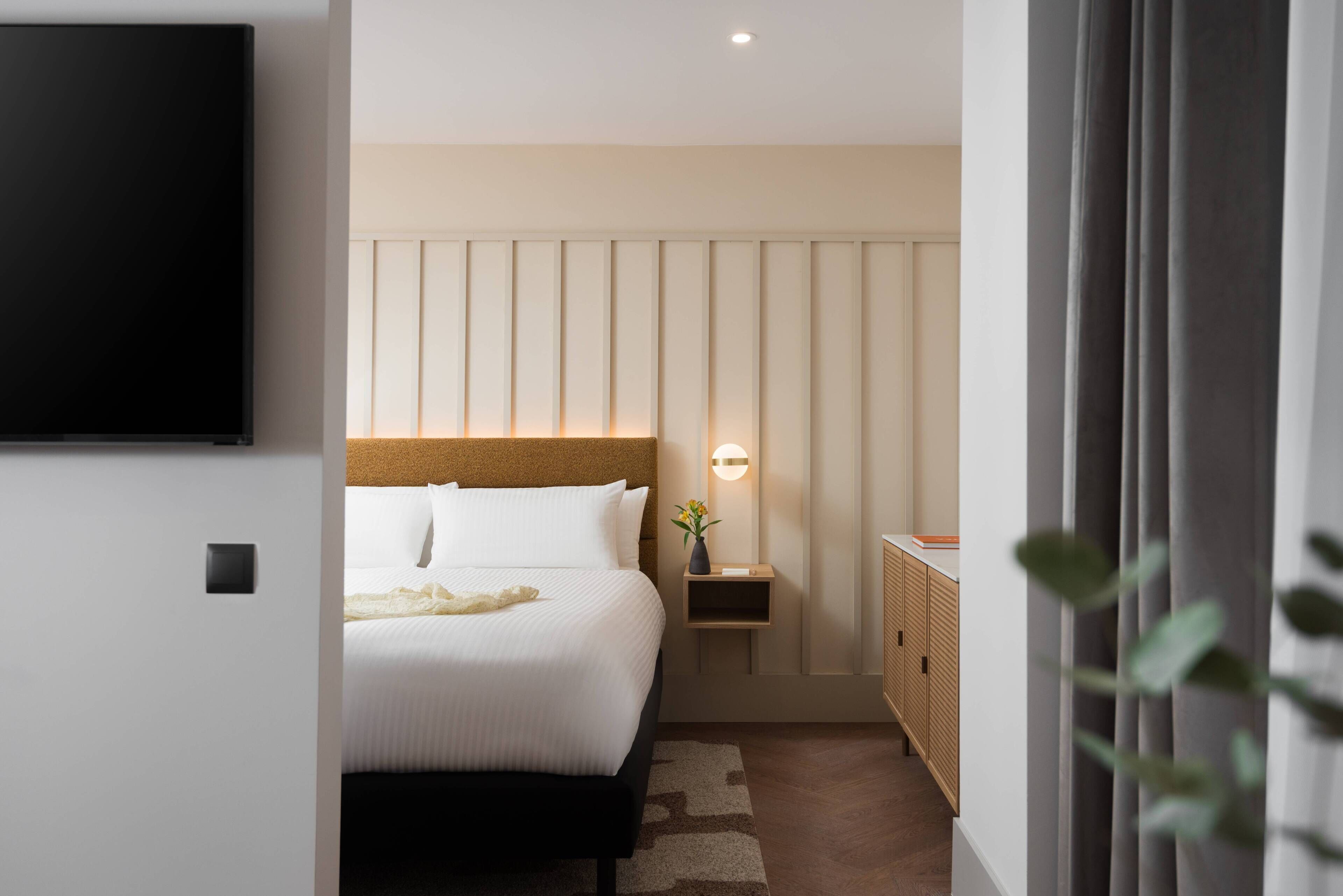 A modern hotel room with a large bed, a bedside table with a lamp, a TV mounted on the wall, and a wardrobe.
