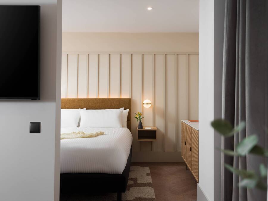 A modern hotel room with a large bed, a bedside table with a lamp, a TV mounted on the wall, and a wardrobe.