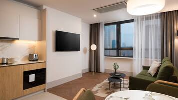 Una habitación de hotel moderna con cocina, una mesa de comedor para dos personas, un sofá verde y un gran ventanal con cortinas.