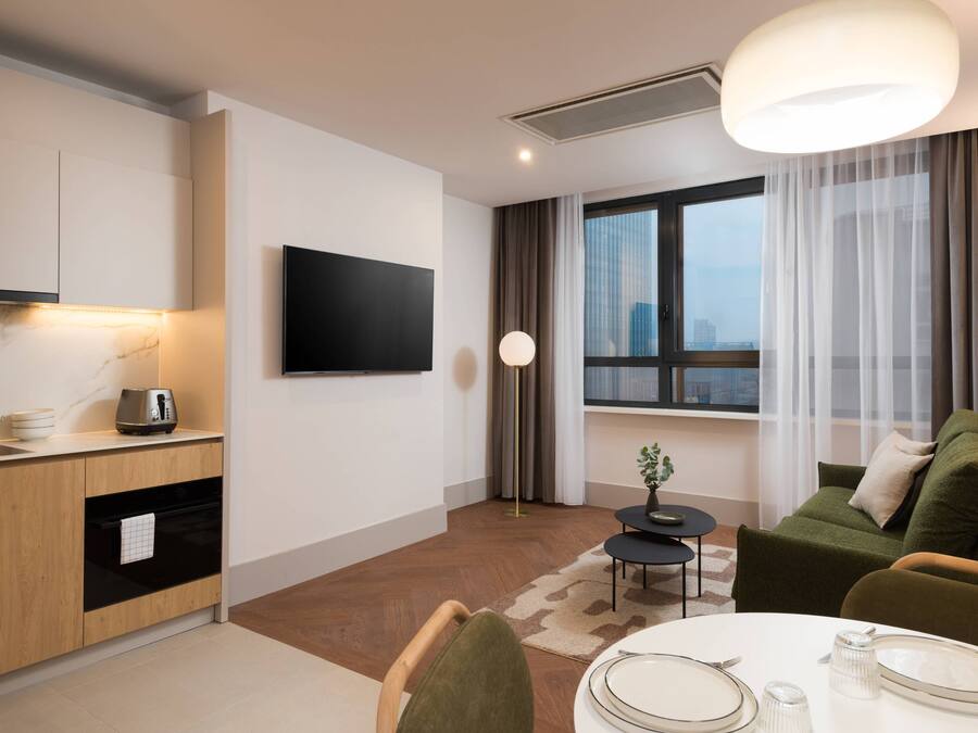 Una habitación de hotel moderna con cocina, una mesa de comedor para dos personas, un sofá verde y un gran ventanal con cortinas.