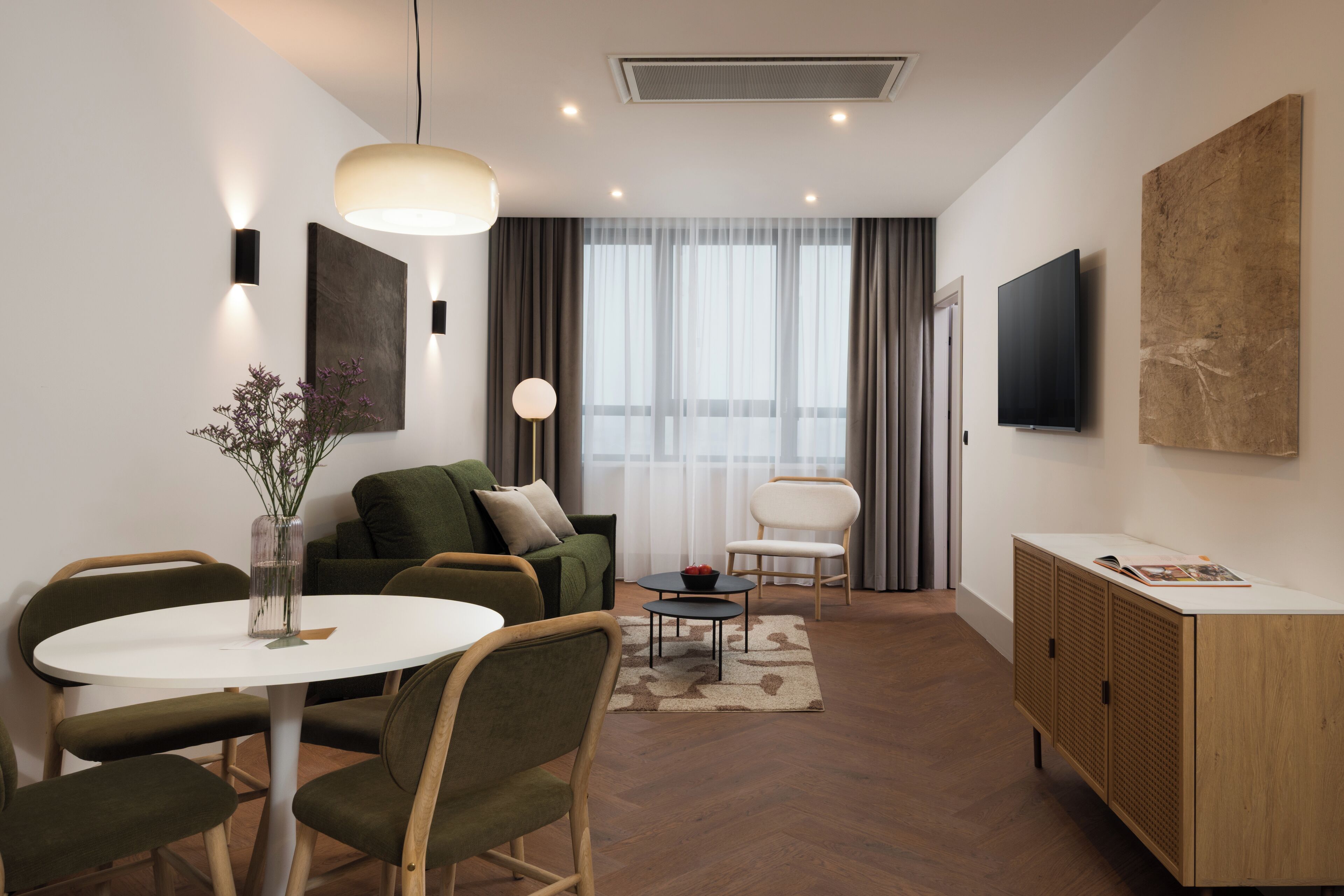 A modern living room with a round dining table, green sofa, and a TV mounted on the wall.