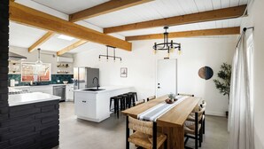 A modern kitchen with white cabinetry, a central island, and a dining area with a wooden table and wicker chairs. - Pool•Hot Tub•Firepit•Fully Fenced with Epic Views (Joshua Tree)