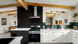 A modern kitchen with a black brick accent wall, stainless steel appliances, white cabinetry, and wooden beams. - Pool•Hot Tub•Firepit•Fully Fenced with Epic Views (Joshua Tree)