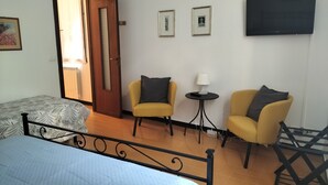 A room with a bed, two yellow armchairs, a small table with a lamp, a TV on the wall, and a door leading to another room. - Gianlu' Cosy Apartment with A/C (Monterosso al Mare)