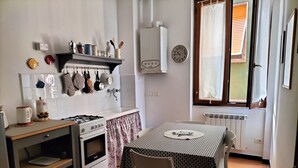 A small kitchen with a gas stove, oven, and a table with a chair. There are hanging utensils and a clock on the wall. - Gianlu' Cosy Apartment with A/C (Monterosso al Mare)