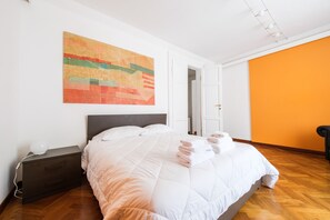 Un lit bien fait, avec une couette blanche et des serviettes pliées, une table de chevet avec une lampe, et un tableau abstrait aux couleurs vives accroché au mur.