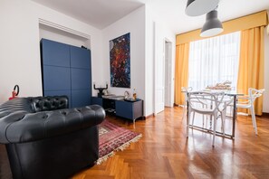 Un salon moderne avec un canapé en cuir noir, un meuble bleu, un coin repas avec des chaises blanches et un grand tableau accroché au mur.