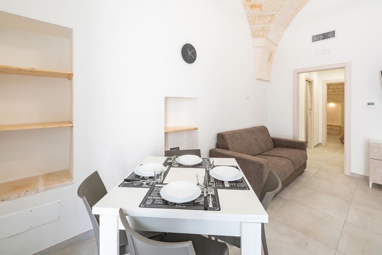 A dining area with a white table set for four, a brown sofa, and a clock on the wall.