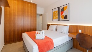 Moderni hotellihuone, jossa on puinen vaatekaappi, valkoisilla lakanoiden ja oranssinvärisen peiton peitetty sänky sekä kaksi abstraktia taideteosta seinällä.
