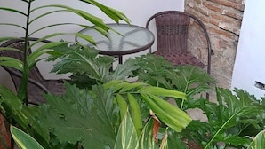 Un petit espace extérieur aménagé avec une table à plateau de verre et des chaises en osier, le tout entouré d’une végétation luxuriante.