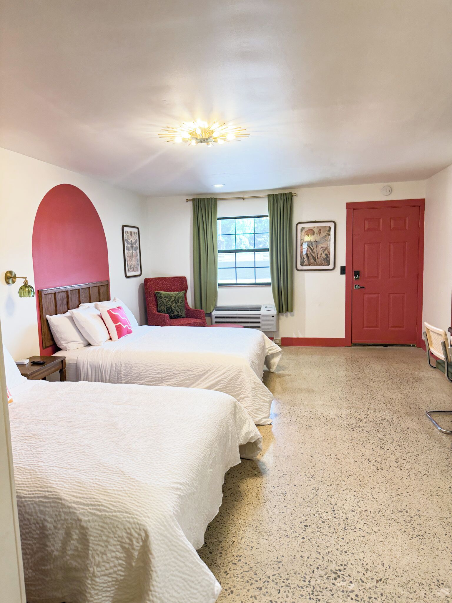 Une chambre d’hôtel avec deux lits, une porte rouge, une fenêtre avec des rideaux verts et un luminaire au plafond.