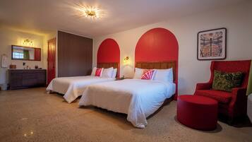 A hotel room with two beds, a red chair, a round ottoman, a vanity with a mirror, and a framed picture on the wall.