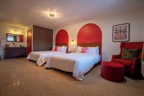 Habitación de hotel con dos camas, una silla roja, un otoman redondo, un tocador con espejo y un cuadro en la pared.
