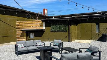 An outdoor seating area with a modern design, featuring a mix of sofas and tables, set against a backdrop of a building with a wooden facade and a clear blue sky.