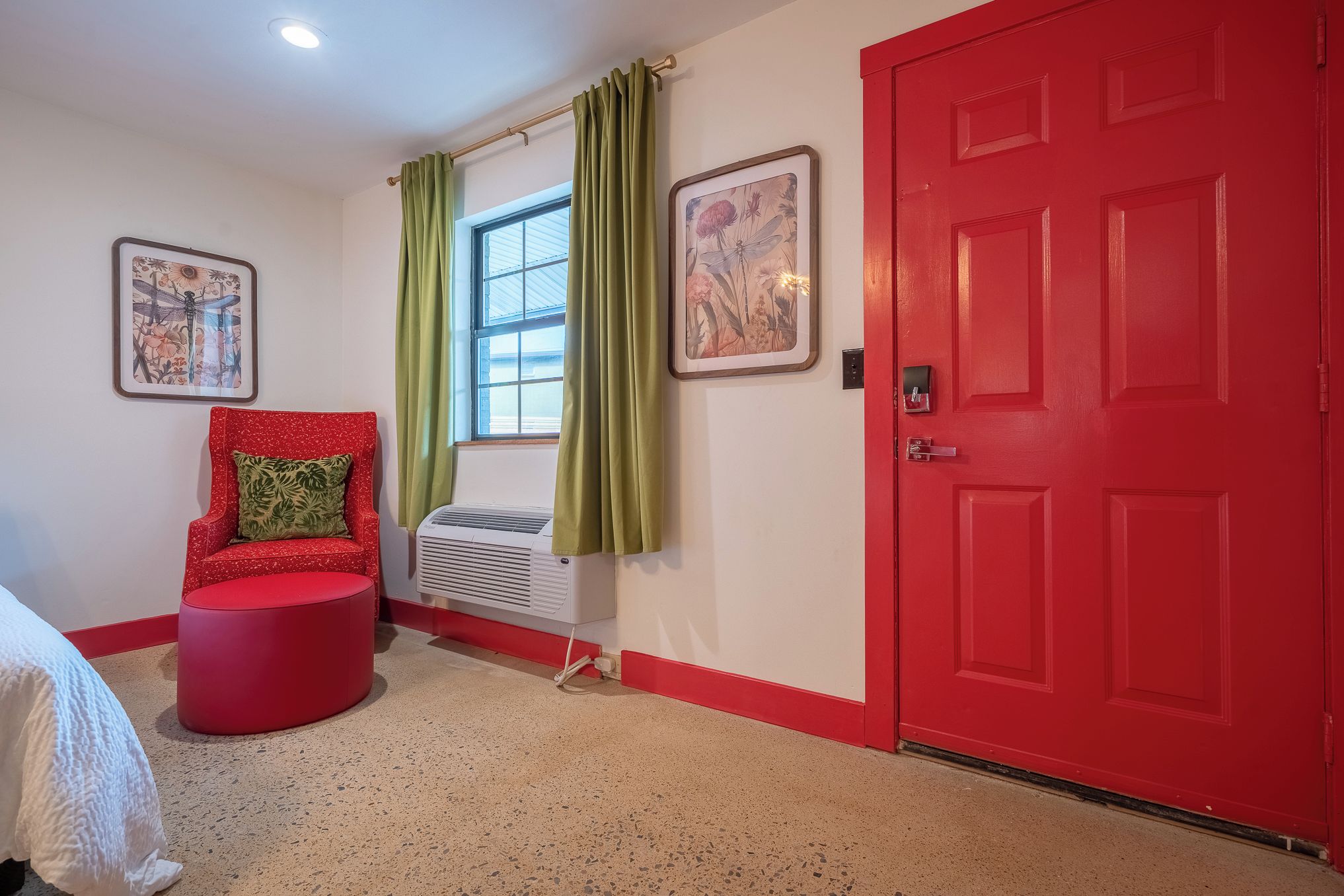 A room with a red door, a red chair with a green cushion, a window with green curtains, and a wall-mounted air conditioner.