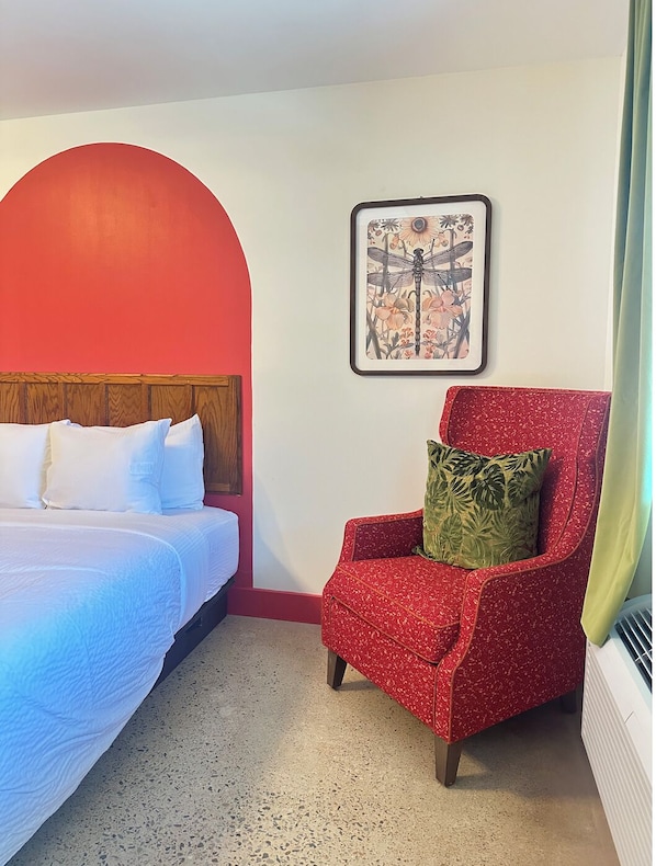 Ett hotellrum med en säng, en röd fåtölj, ett inramat konstverk och en röd valvöppning.
