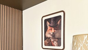 A neatly made bed with white linens, a wooden headboard, a framed picture of a fox on the wall, and a lamp with a tree pattern on the bedside table.