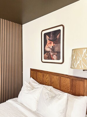 Una cama bien hecha con sábanas blancas, un cabecero de madera, un cuadro enmarcado de un zorro en la pared y una lámpara con un estampado de árbol en la mesita de noche.