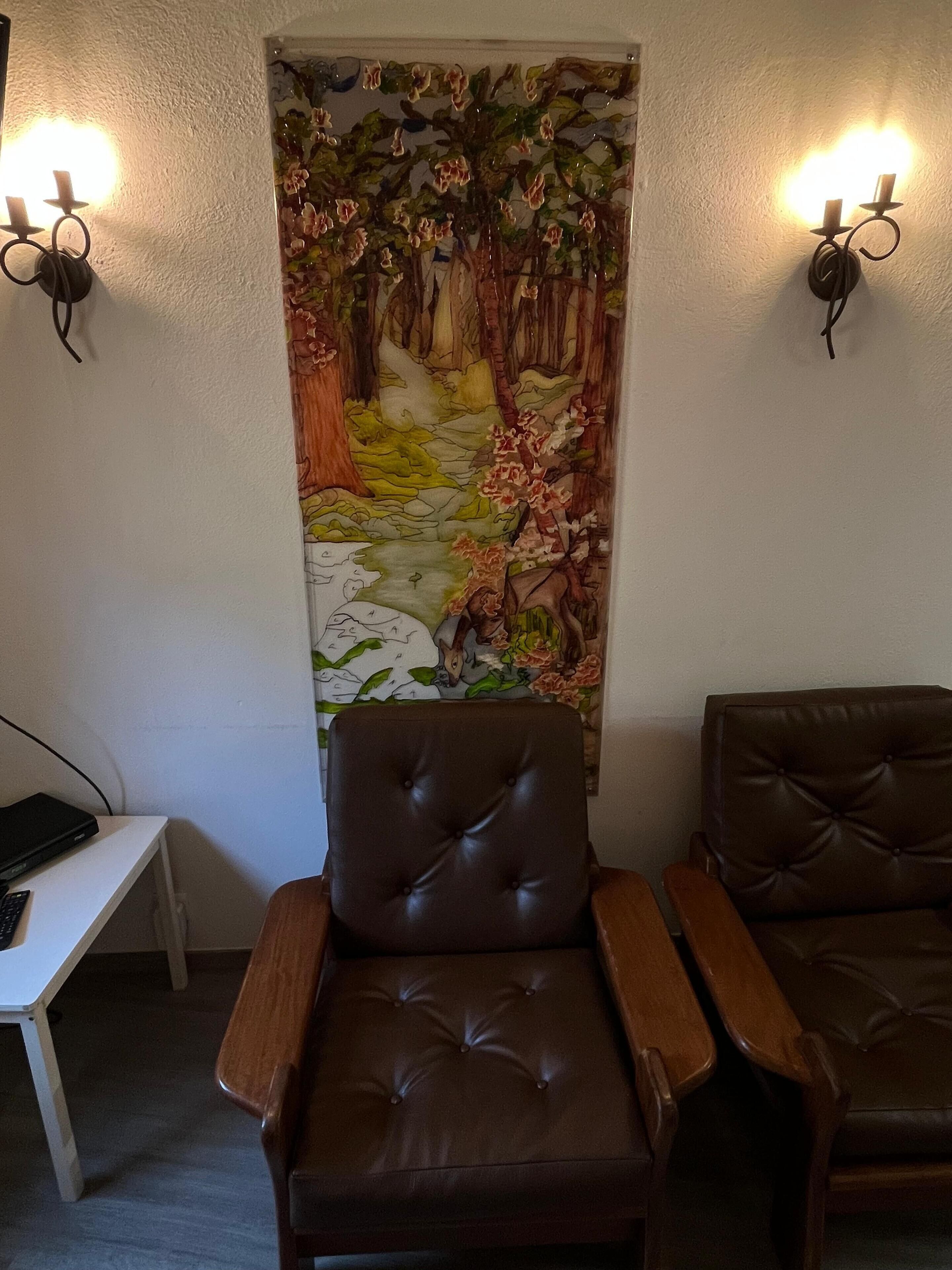Quarto com uma grande tapeçaria com tema de floresta na parede, duas poltronas de couro e uma pequena mesa com um laptop.