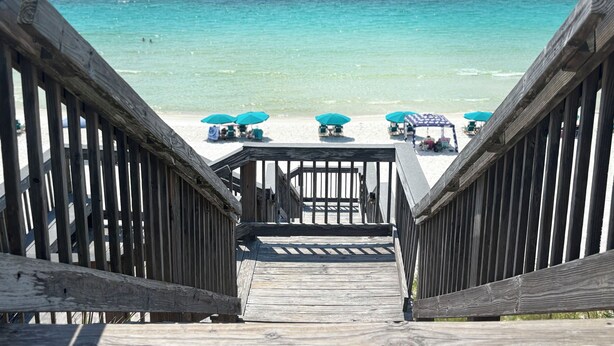 Un escalier en bois menant à une plage aux eaux turquoise, bordée de parasols.