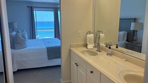 Une chambre d’hôtel avec un lit, une salle de bain comprenant un lavabo et un miroir, et une vue sur la mer depuis la fenêtre.