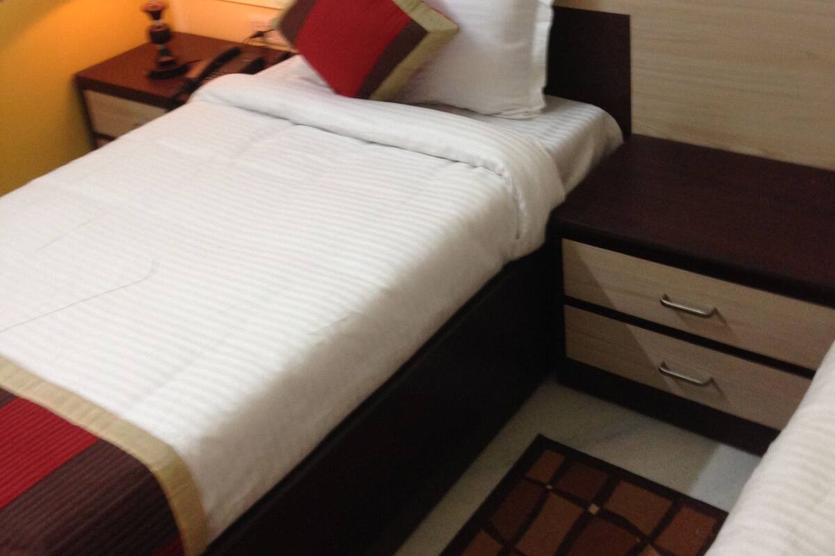 A single bed with a white comforter and two pillows, a bedside table with a lamp, and a small drawer unit.