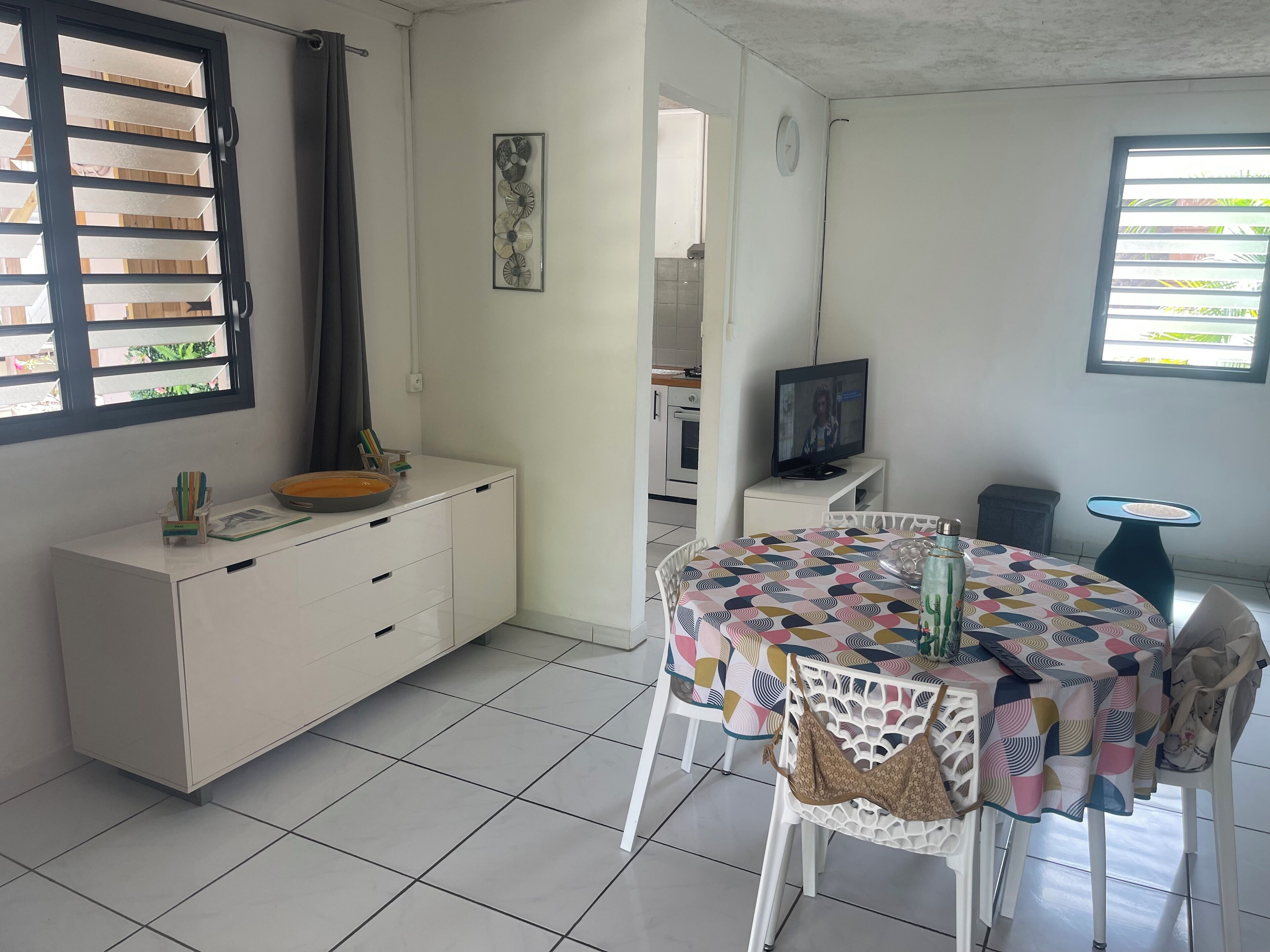 A modern kitchen with white cabinetry, a dining area with a colorful tablecloth, and a television on a stand.