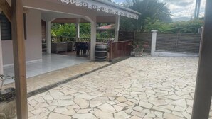 A covered patio area with stone flooring, outdoor seating, and a view of the surrounding greenery. - Country house 20 min from the beach (Rivière-Pilote)
