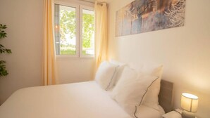 Cama bem arrumada com lençóis e almofadas brancas, uma luminária de cabeceira, uma janela com cortinas e um quadro abstrato fixado na parede.