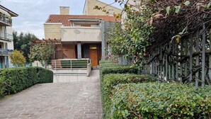 A paved pathway leading to a modern building with a tiled roof, surrounded by well-maintained hedges and a metal fence.