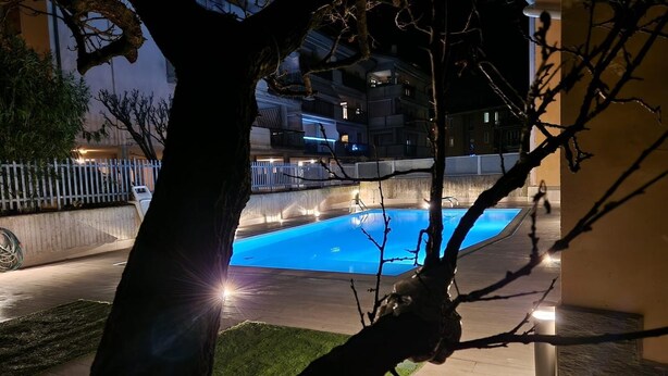 A nighttime view of a swimming pool area with a tree in the foreground, a fence, and a building in the background.