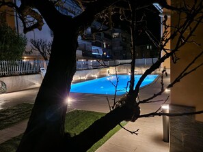A nighttime view of a swimming pool area with a tree in the foreground, a fence, and a building in the background. - iris b&b (Montorio)