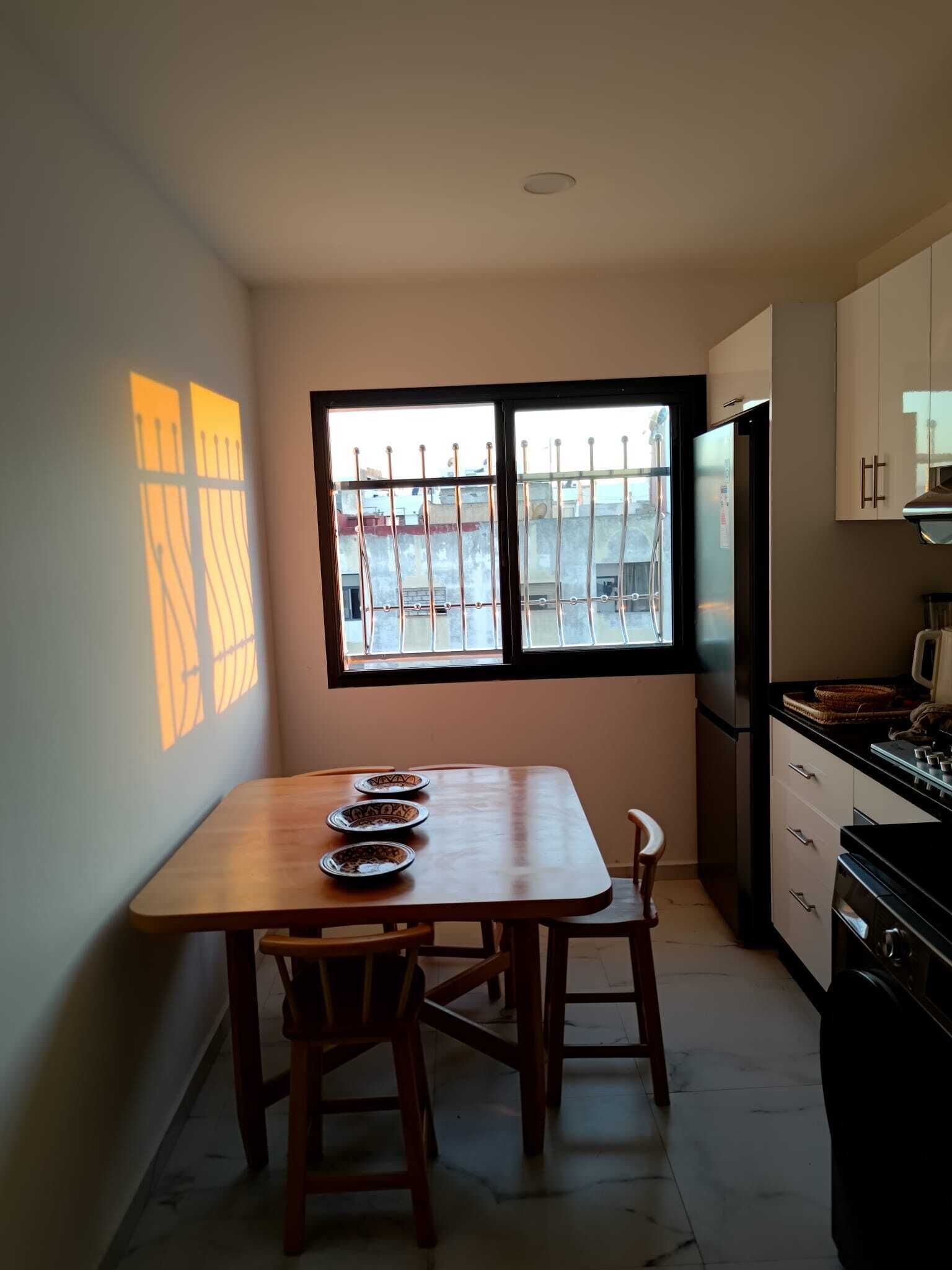 A small kitchen with a dining table and chairs, a refrigerator, and a window with a view of buildings outside.