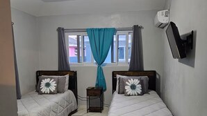 A small, clean room with two beds, a ceiling fan, an air conditioning unit, and a window with curtains. - Queen's Court 2-bedroom apartment in Nassau! (Nassau)