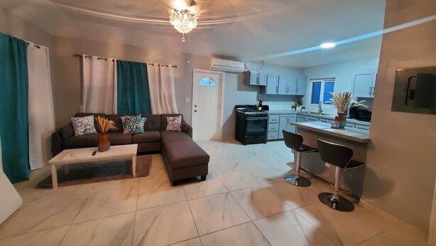 A modern living room with a sectional sofa, a coffee table, and a dining area with two bar stools. A kitchen with a stove and cabinets is visible in the background. - Queen's Court 2-bedroom apartment in Nassau! (Nassau)