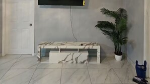 A living room with a flat-screen TV mounted on the wall, a marble bench, a ceiling fan, and a potted plant. - Queen's Court 2-bedroom apartment in Nassau! (Nassau)