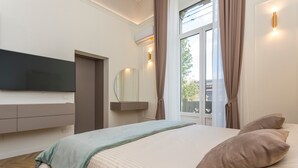 Ein modernes Schlafzimmer mit einem Flachbildfernseher, einem Bett mit einer beige-grünen Decke, einem großen Fenster mit Vorhängen und einer wandmontierten Klimaanlage.