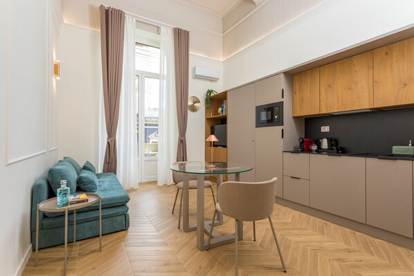 A compact living space with a sofa, a glass-top dining table, a kitchen area with wooden cabinets, and a herringbone-patterned floor.
