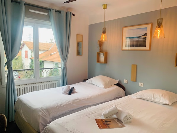 Une chambre d’hôtel avec deux lits, une fenêtre avec des rideaux, un radiateur, un tableau encadré au mur et un luminaire suspendu.