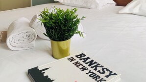 Un lit bien fait, avec des draps blancs, une plante verte, une serviette enroulée et un livre intitulé « BANKSY À NEW YORK » posé sur le lit.