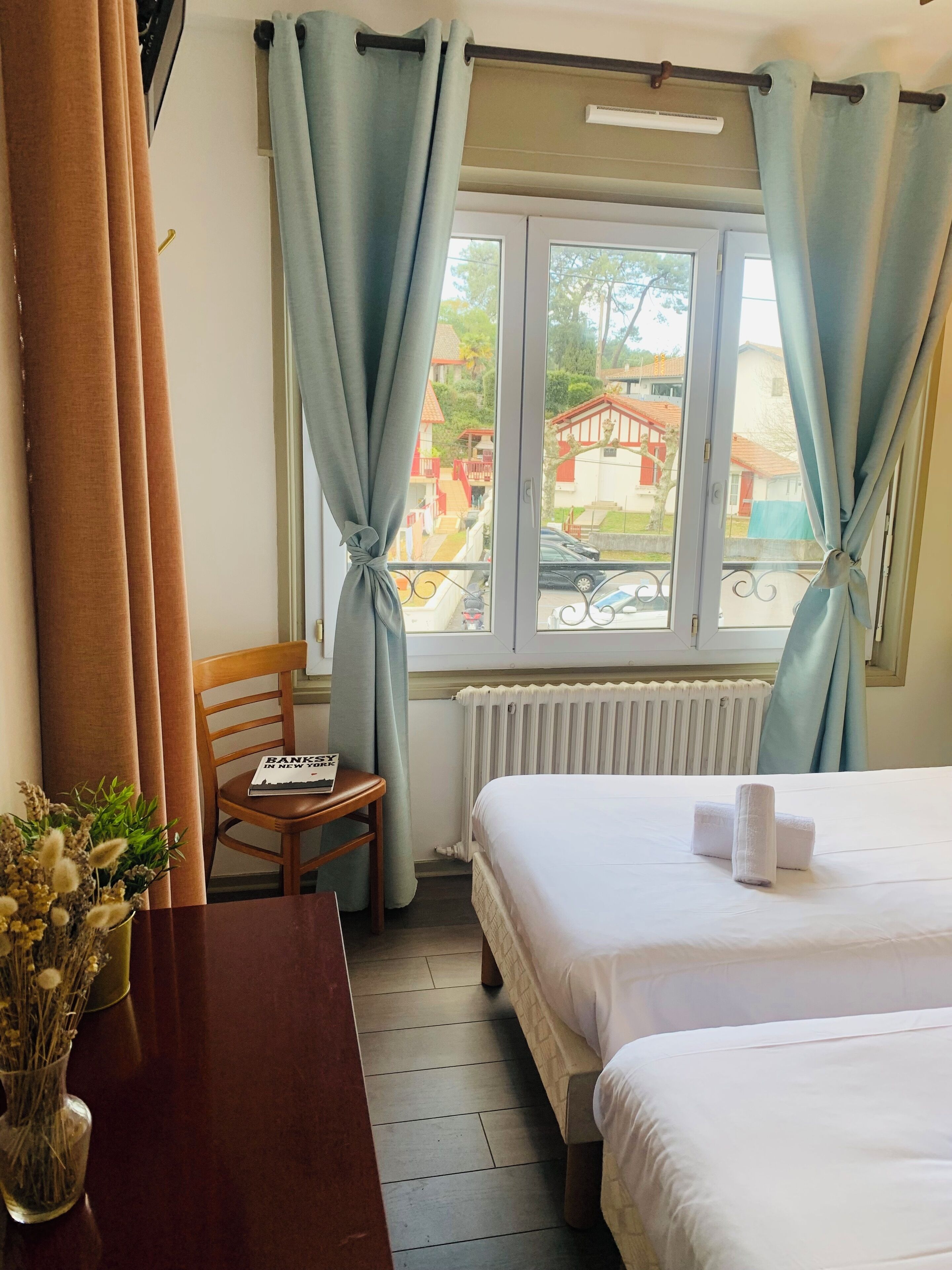 Een hotelkamer met twee bedden, een houten stoel, een tafel met een boek en een raam met gordijnen.
