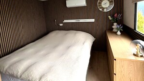 Een knus slaapkamer met een bed, een houten bureau, een wandairconditioner, een klok en een raam met uitzicht.