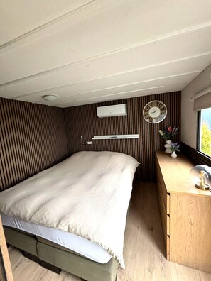 A compact bedroom with a bed, a wooden desk, a wall-mounted air conditioner, a clock, and a window with a view.