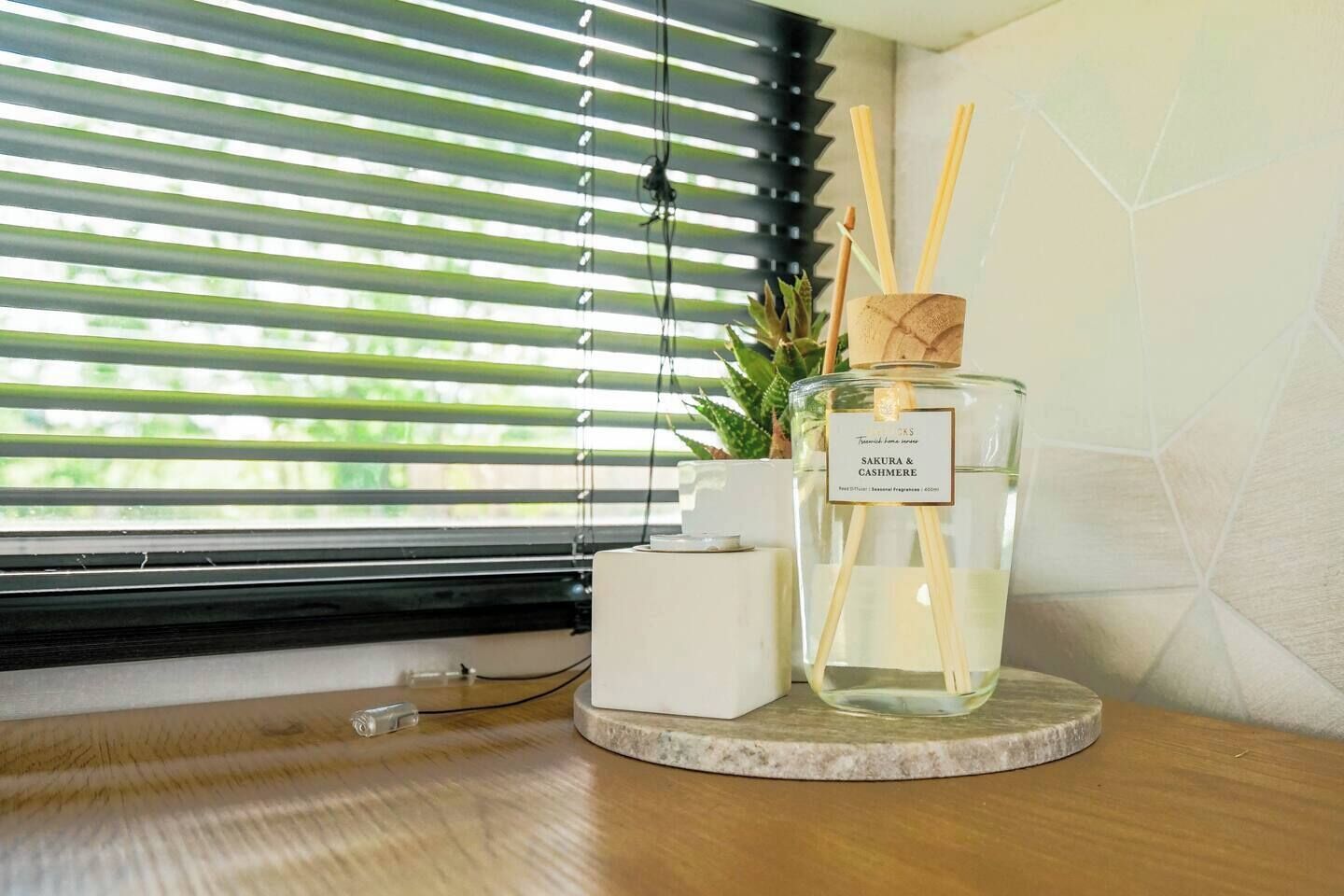 A bottle of Sakura & Cashmere fragrance on a marble coaster, a small potted plant, and a white candle on a wooden surface by a window with blinds.