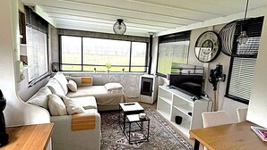 Een moderne woonkamer met een bank, een salontafel en een televisie. Er zijn grote ramen en een kamerkleed met een patroon.