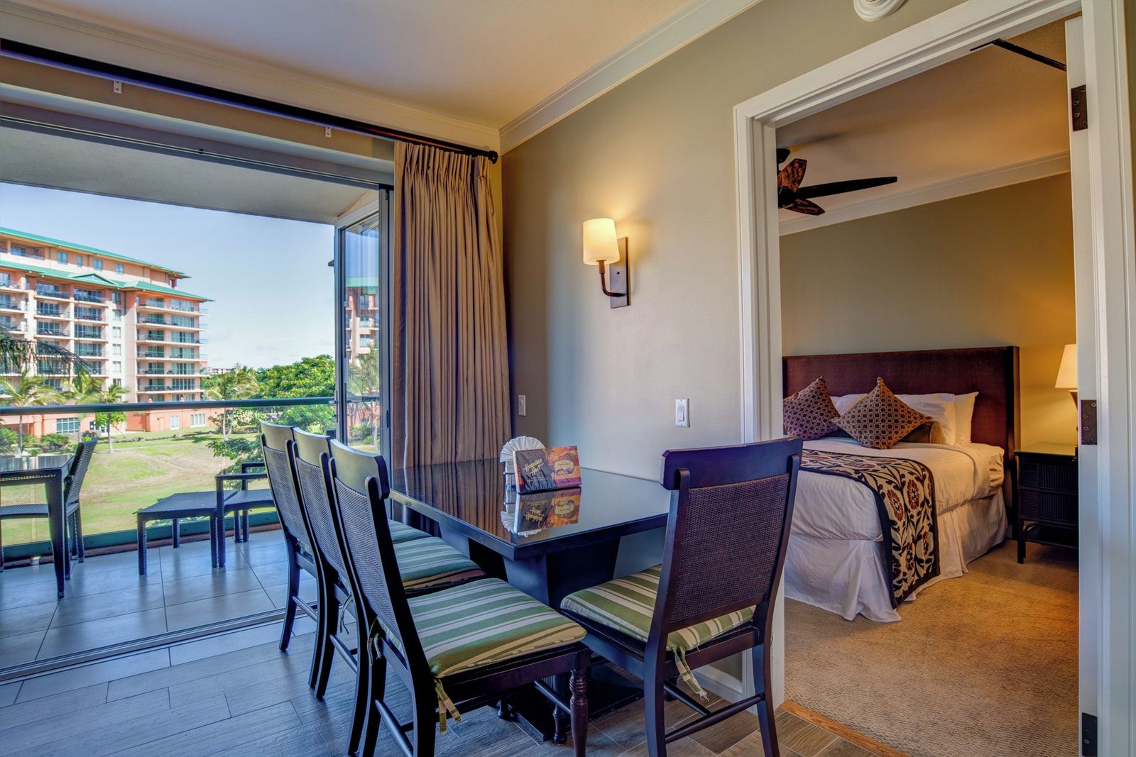 Une chambre d’hôtel avec un balcon, un lit avec une tête de lit, une table avec des chaises et une vue sur un bâtiment et la verdure environnante.