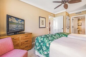 Een hotelkamer met een televisie, een bed met een sprei met bladprint, een houten kast, een plafondventilator en een deur naar een andere kamer.