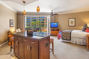 A hotel room with a kitchenette, a TV, a bed with patterned bedding, a window with a view of a building, and a ceiling fan.