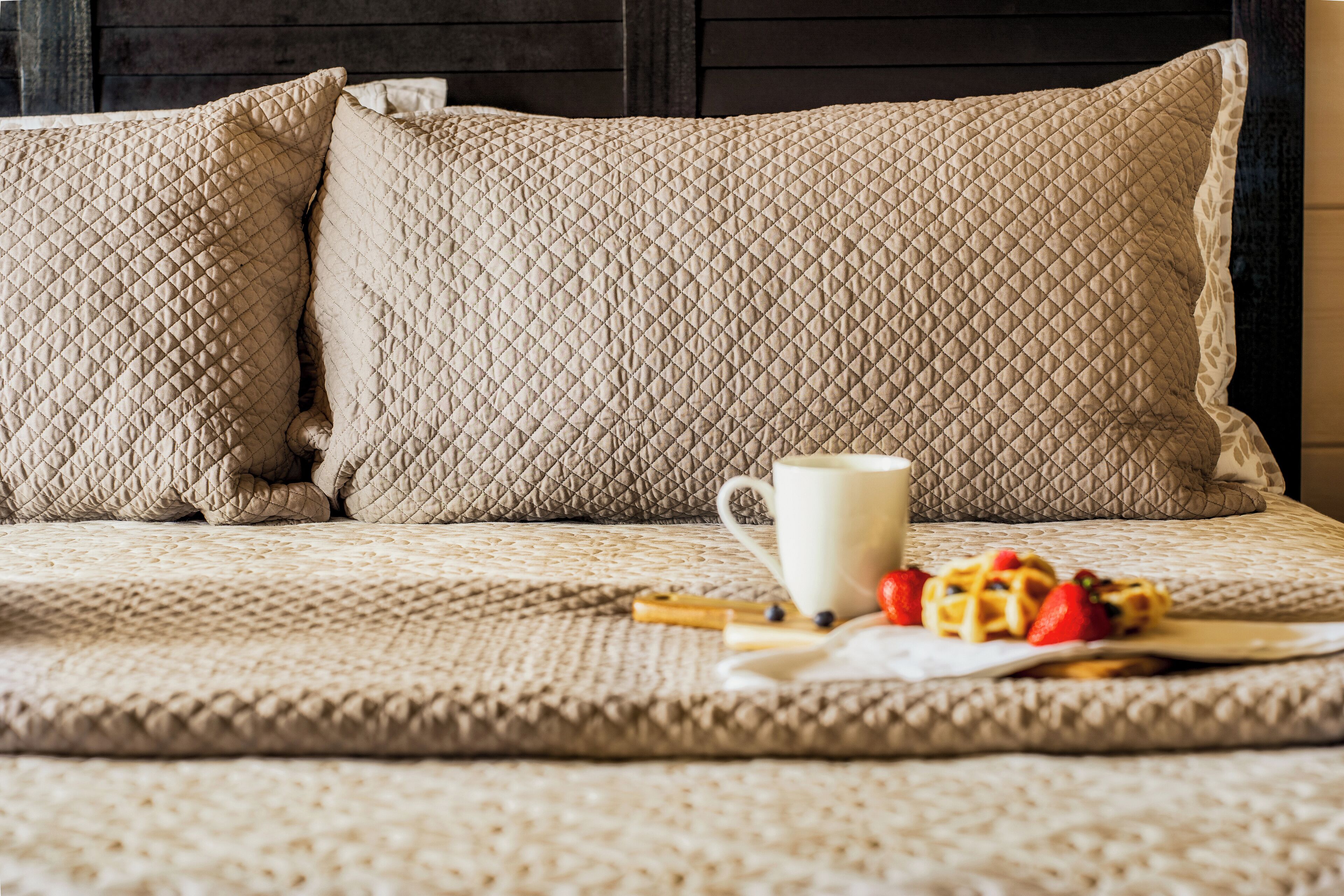 Un lit recouvert d’une couverture matelassée beige, deux oreillers, une tasse blanche et une assiette de gaufres aux fraises.